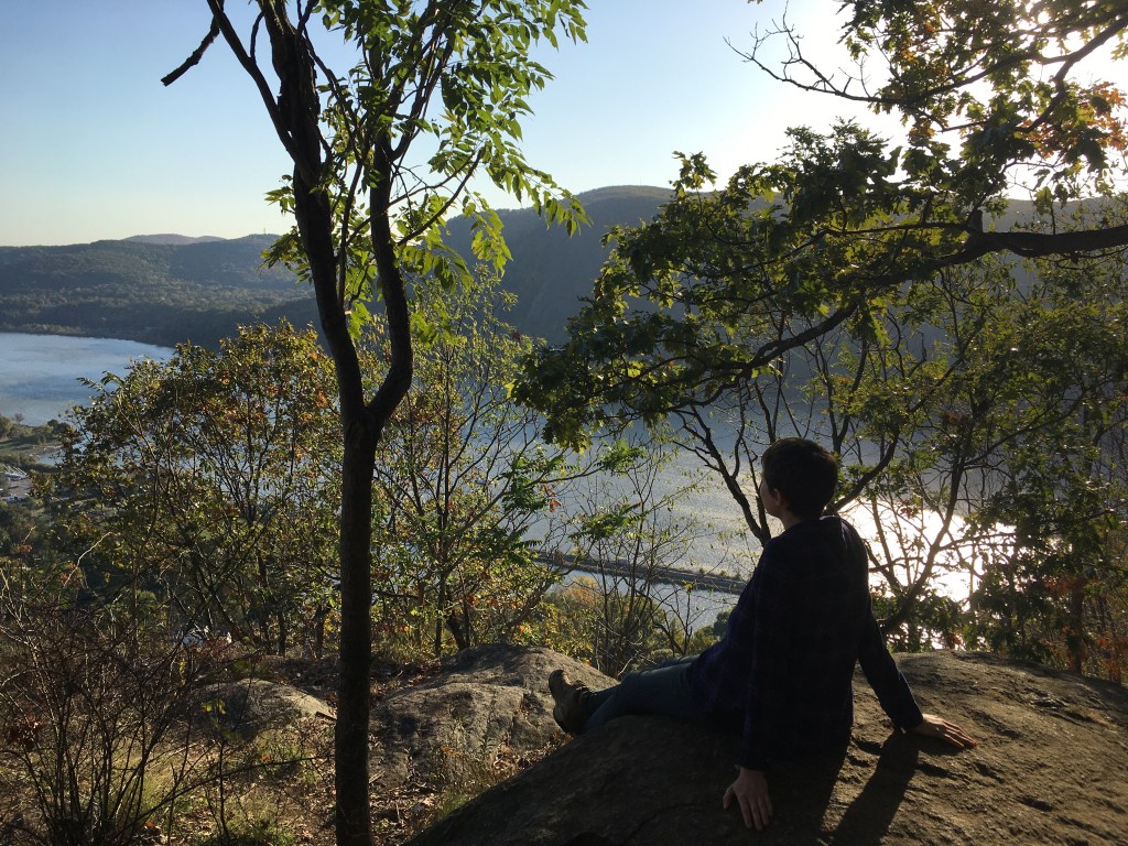 A person sitting on a large rock, facing away from the camera and toward a distant lake with a large hill behind it